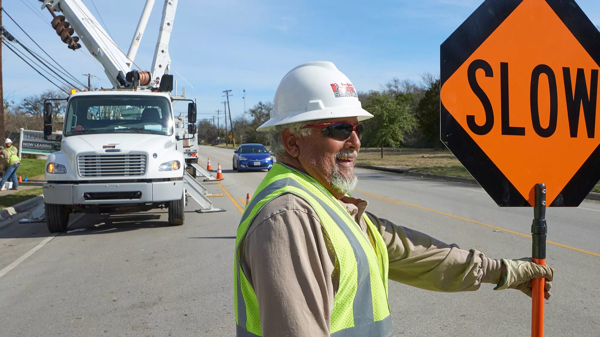 A smiling man in a Linetec hard hat holds a slow traffic sign on the road.